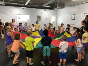 Children stand holding a big parachute listening to the teachers explain the next fun activity.